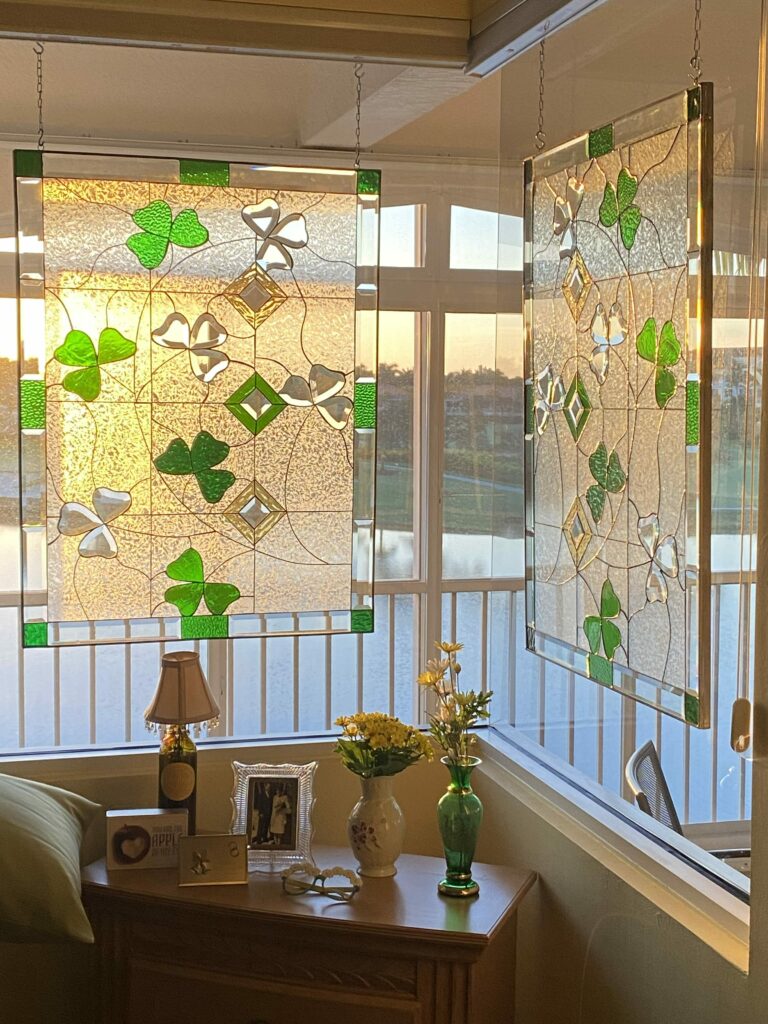 Two rectangular stained glass panels, each featuring a repeating pattern of vibrant green shamrocks, clear textured glass, and sparkling clear beveled diamond and clover shapes, hang suspended by chains in a sunlit corner window. The warm evening light shines through the panels, revealing an outdoor scene of a lake and lush greenery.