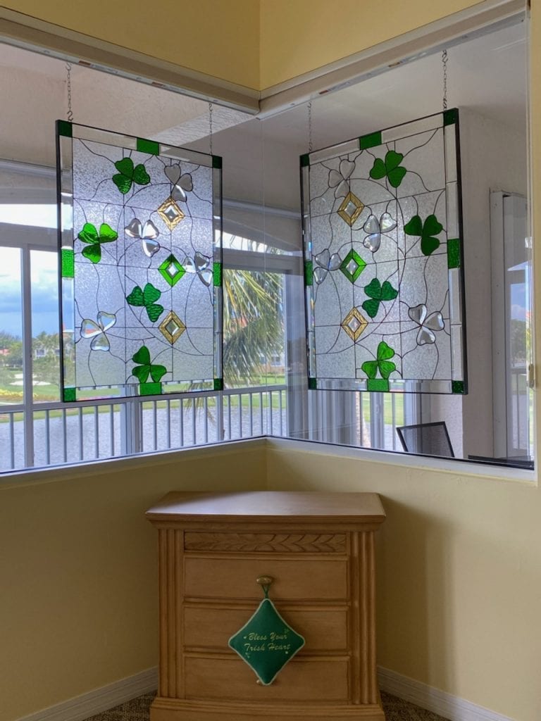 Two rectangular, suspended leaded stained glass panels feature a design of green and clear textured shamrocks, interspersed with clear textured glass and gold beveled diamond accents, hanging in front of large windows with a view of an outdoor waterway and palm trees. A wooden dresser sits beneath them with a decorative green pillow.