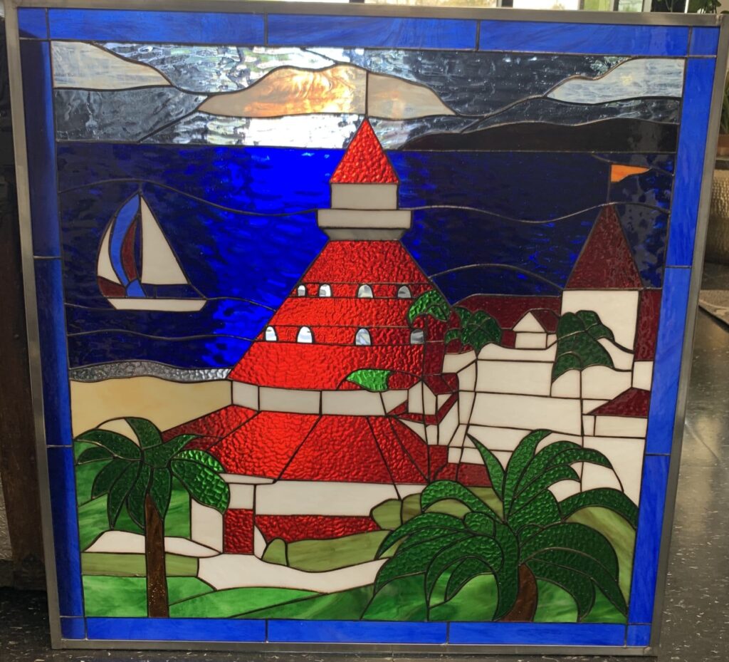 A square stained glass panel featuring a vibrant coastal scene. A prominent building with a textured red roof and white walls, resembling a resort or lighthouse, stands central. Green palm trees are scattered in the foreground and around the building. A white sailboat with blue and red details sails on deep blue, wavy water. A narrow strip of sandy beach separates the water from the land. The sky above includes clear and cloudy glass pieces. The panel has a silver-colored metal frame.
