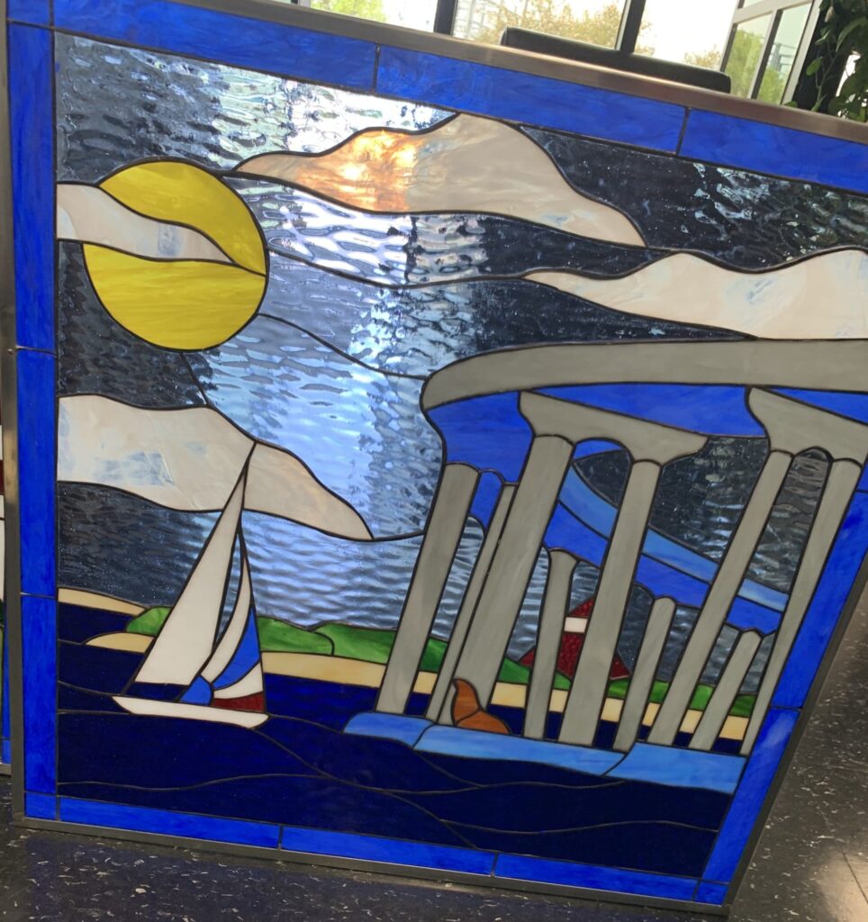 A rectangular stained glass art panel in a metal frame, featuring a vibrant coastal scene. A bright yellow sun and flowing white clouds fill a textured blue sky. Below, a white sailboat with blue details glides across deep blue water. A sandy shoreline with green hills leads to a gray pier or bridge supported by columns, under which a reddish-brown seal rests. The entire panel is bordered by a rich royal blue glass frame.