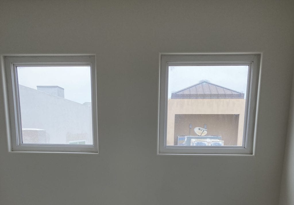 Two rectangular clear glass windows, framed in white, set into a light-colored interior wall, revealing distant building rooftops, an AC unit, and a satellite dish under a bright, overcast sky.