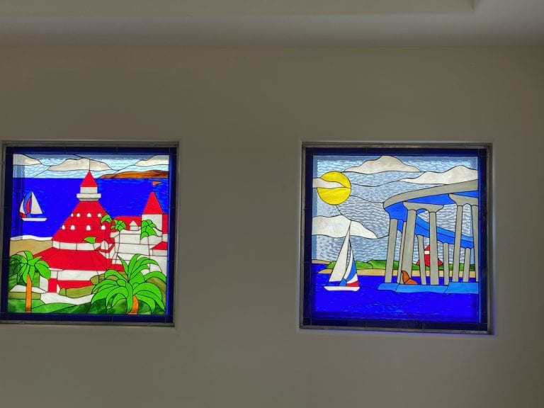Two rectangular stained glass window panels depicting iconic San Diego coastal scenes. The left panel shows a red-roofed hotel, palm trees, and a sailboat on the blue ocean. The right panel features a large bridge, a sailboat, a lighthouse, and a bright yellow sun over the ocean.