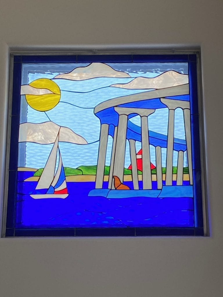 A vibrant rectangular stained glass window panel depicting a coastal scene. A bright yellow sun is in the upper left, surrounded by white clouds in a light blue sky. A white sailboat with blue and red accents sails on a deep royal blue sea. On the right, tall grey columns with blue caps rise from the water, with a red lighthouse-like building and green land visible behind them. The entire panel has a dark blue border.