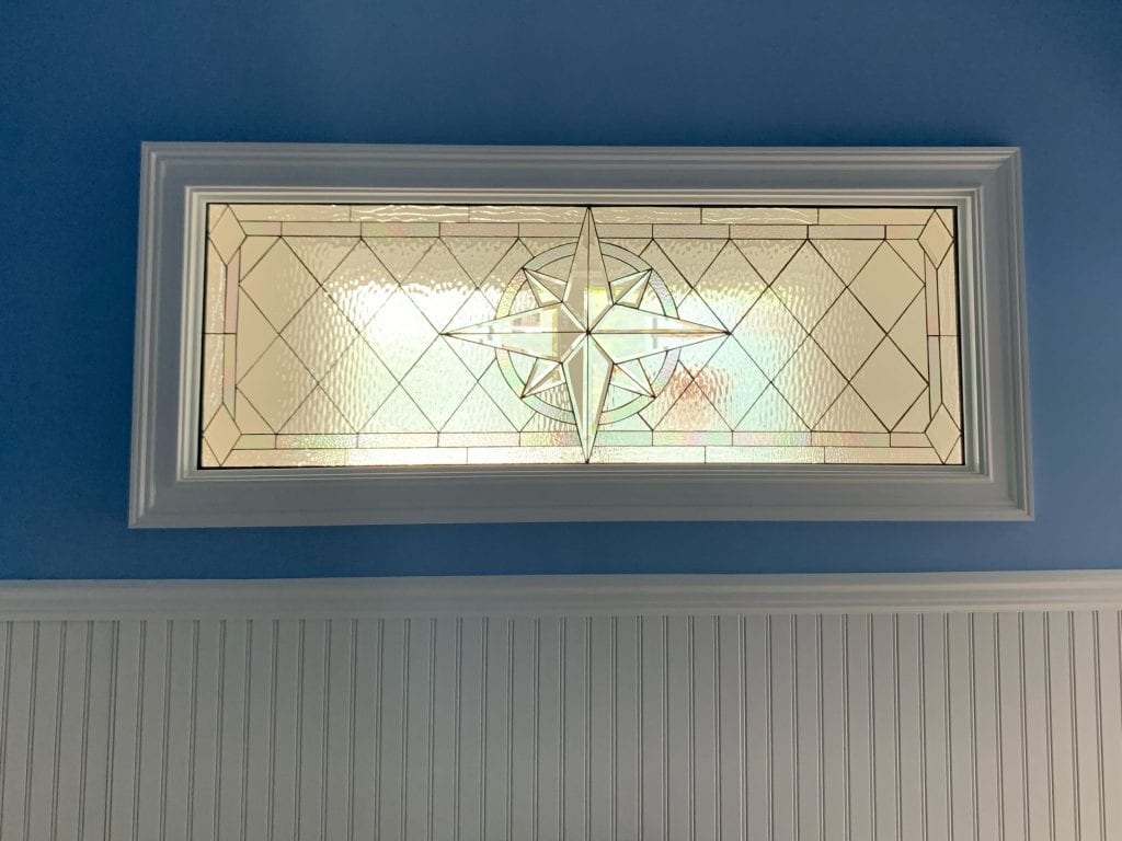 A rectangular clear leaded stained glass transom panel with a prominent central compass rose design, surrounded by a geometric pattern of clear textured and beveled glass sections, set in a white frame against a blue wall above white beadboard wainscoting.