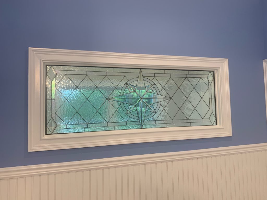 A rectangular clear leaded stained glass transom window, set in a white frame against a blue wall with white wainscoting. The panel features a central compass rose motif made of clear beveled glass, surrounded by a geometric pattern of clear textured glass diamonds and rectangles.