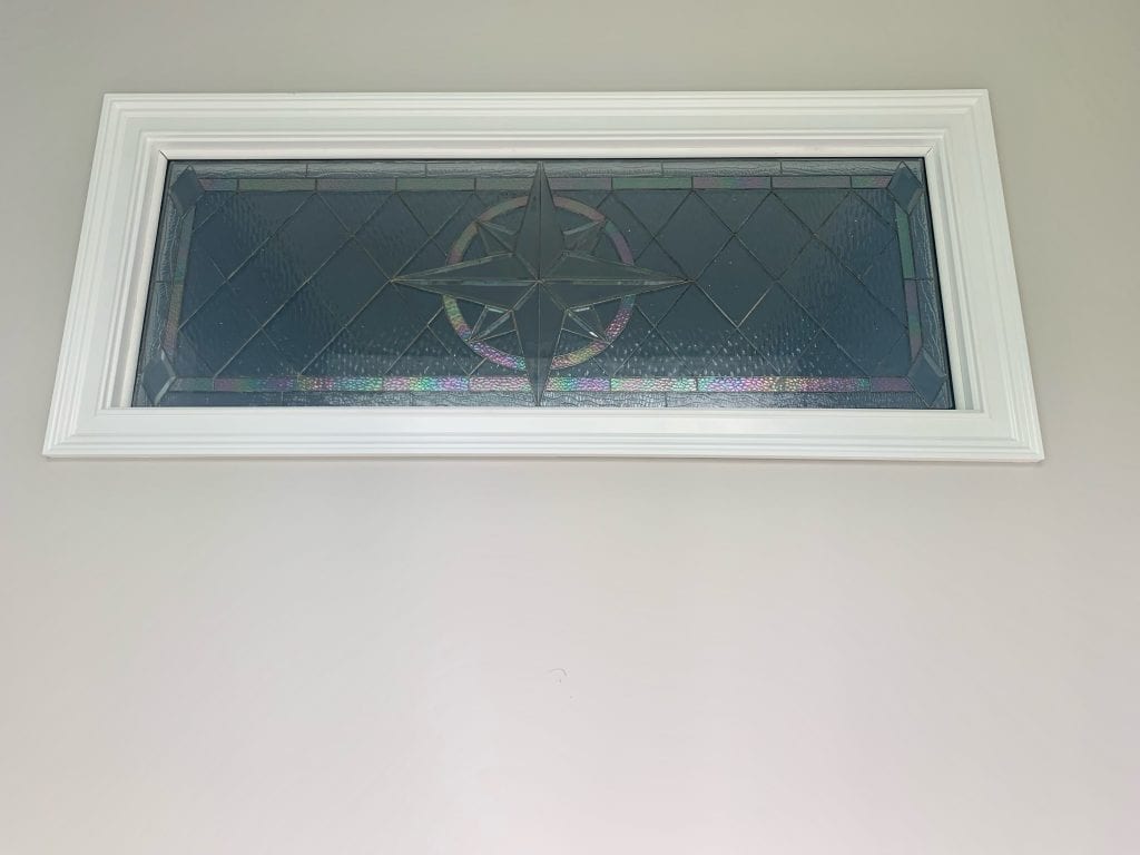 A rectangular transom window with a white frame, featuring a leaded stained glass panel. The panel displays a clear beveled compass rose design at its center, surrounded by textured clear glass in a diamond pattern. Iridescent accents are visible within the compass rose and along the bottom border of the panel.