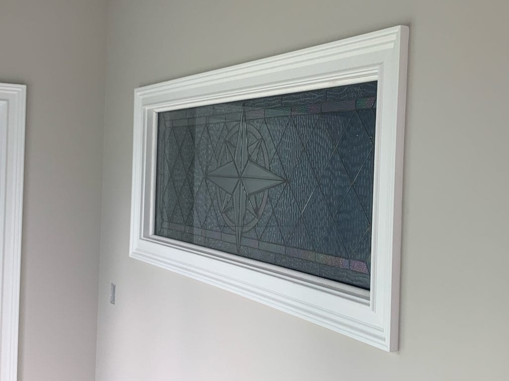 A rectangular transom window featuring a leaded clear stained glass panel. The panel showcases a central beveled starburst or compass rose medallion, surrounded by clear, heavily textured (like waterglass) diamond-patterned glass, all encased in a simple white frame mounted on a light gray wall.