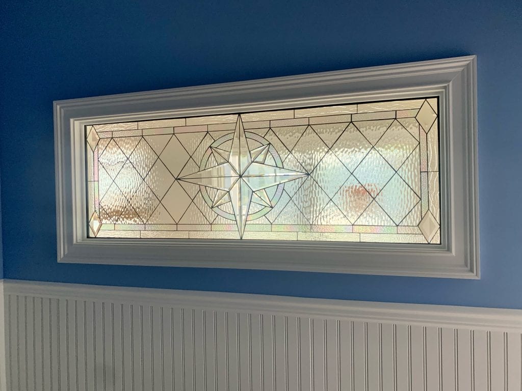 A rectangular, clear leaded stained glass panel with a central compass rose design, surrounded by geometric patterns of clear textured and beveled glass. The panel is installed in a white window frame, against a vibrant blue wall with white beadboard wainscoting below.