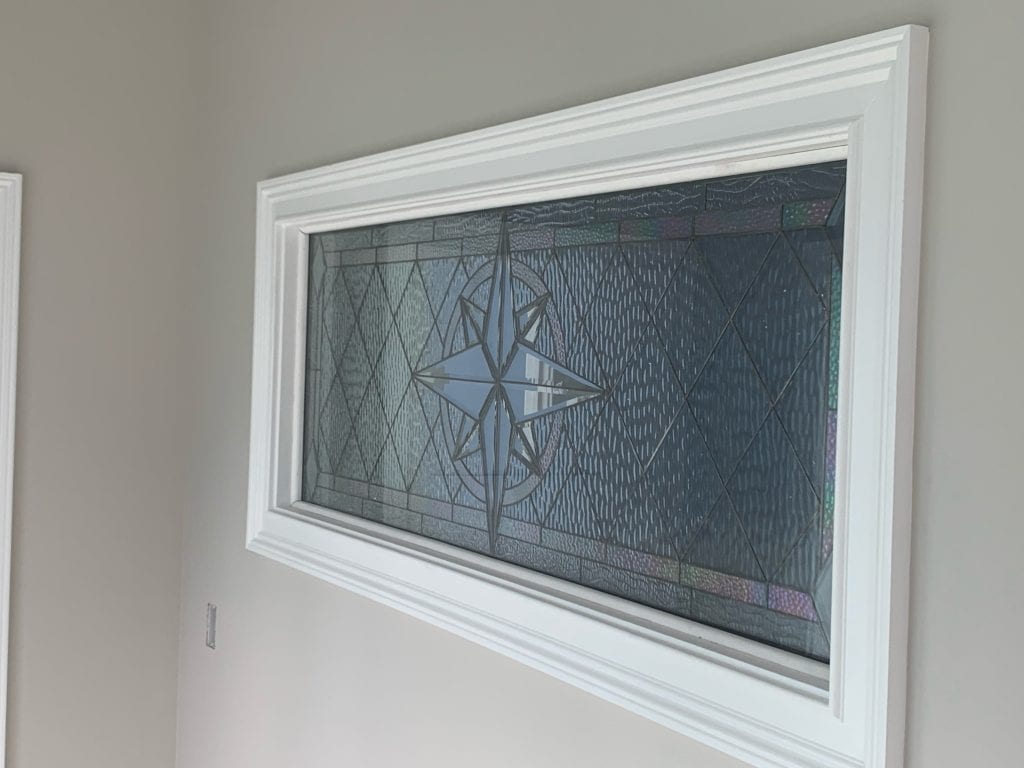 A rectangular leaded stained glass window panel, showcasing a prominent central clear beveled star or compass rose design. The surrounding glass consists of clear, textured, and subtly iridescent sections, arranged in geometric diamond and rectangular patterns. The panel is set within a crisp white frame against a neutral light gray wall.