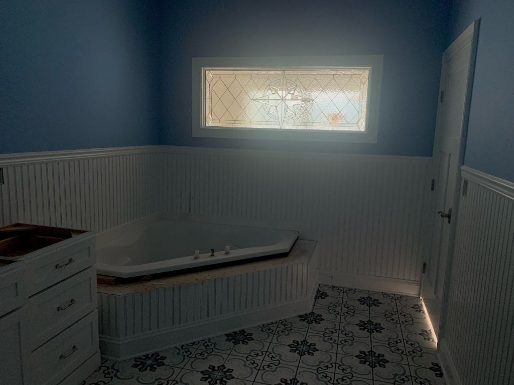 A rectangular clear beveled and textured stained glass window featuring a central starburst design with diamond patterns, installed above a white corner bathtub in a bathroom with deep blue walls, white beadboard wainscoting, and ornate grey and blue patterned floor tiles.