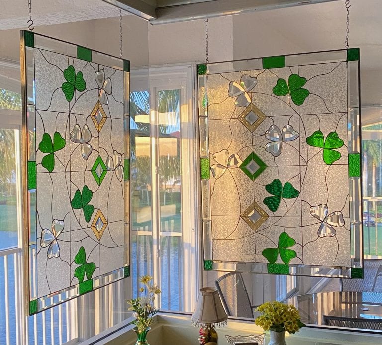 Two rectangular stained glass panels hanging in a sunny window, showcasing a pattern of vibrant green shamrocks, clear textured glass, and shimmering clear beveled diamond and heart-shaped accents. The panels diffuse light and offer privacy.
