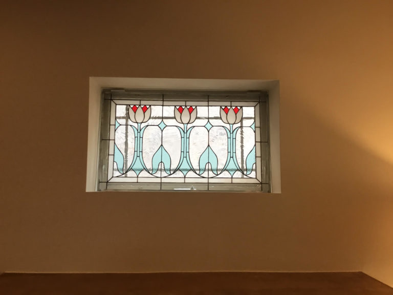 A rectangular leaded stained glass transom window featuring three stylized white tulips with red centers and light blue leaves, set against clear textured glass within a white window frame on a warm brown wall.