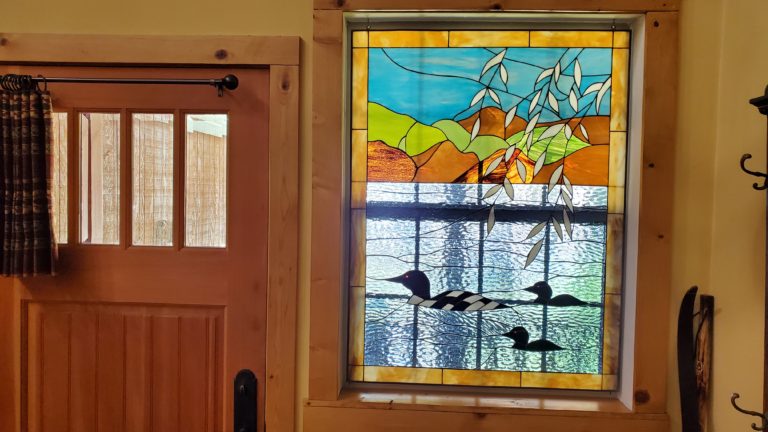 A rectangular stained glass window panel depicting a picturesque lake and mountain landscape. The upper portion features a bright blue sky with light green and earthy brown rolling hills, partially obscured by cascading white and light green willow branches. The lower portion portrays a body of water made of clear, textured rippled glass, where three loons are swimming. The prominent loon in the foreground has a black head, red eye, and a distinctive black and white checkered back. Two smaller, silhouetted black loons with red eyes follow behind it.