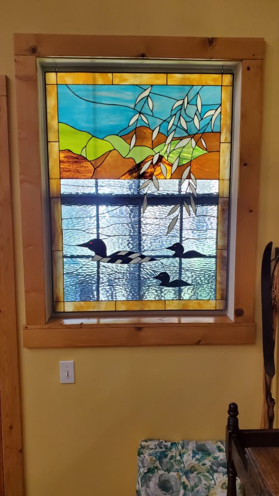 A rectangular stained glass window panel depicting a serene lake and mountain landscape with three loons swimming. The upper section features a blue sky over green and brown rolling hills, with delicate white branches extending from the top. The lower section showcases textured blue glass representing shimmering water, where two adult black loons with red eyes and white checkered patterns, along with a smaller loon chick, gracefully glide. The entire panel is encased in a natural light wood frame.