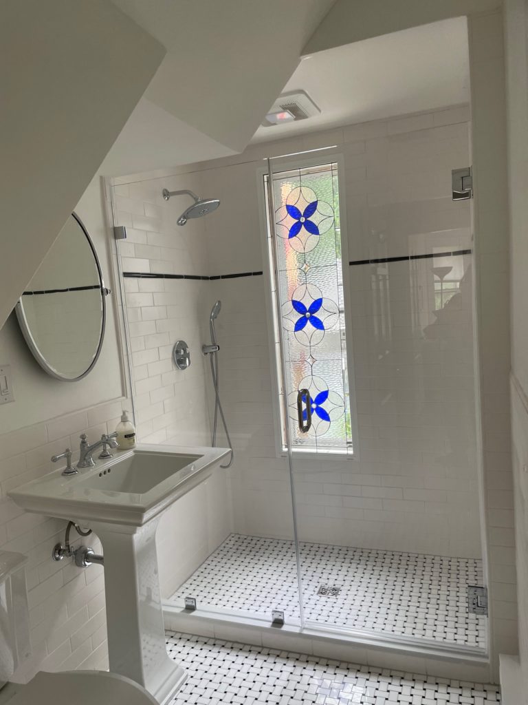 A tall, narrow stained glass window in a modern white tiled shower, featuring three vibrant blue four-petal geometric shapes set within clear textured leaded glass, providing privacy while allowing light through. The shower also has a clear glass enclosure, a white pedestal sink, and black and white basketweave floor tiles.