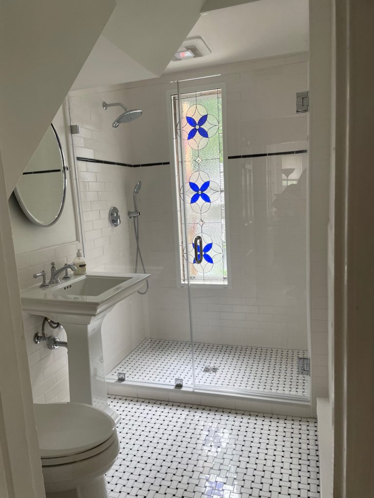 A tall, rectangular stained glass panel with a repeating blue four-petal geometric pattern set against clear, textured glass, serving as a privacy window in a modern bathroom shower with white subway tiles and a black and white mosaic floor.