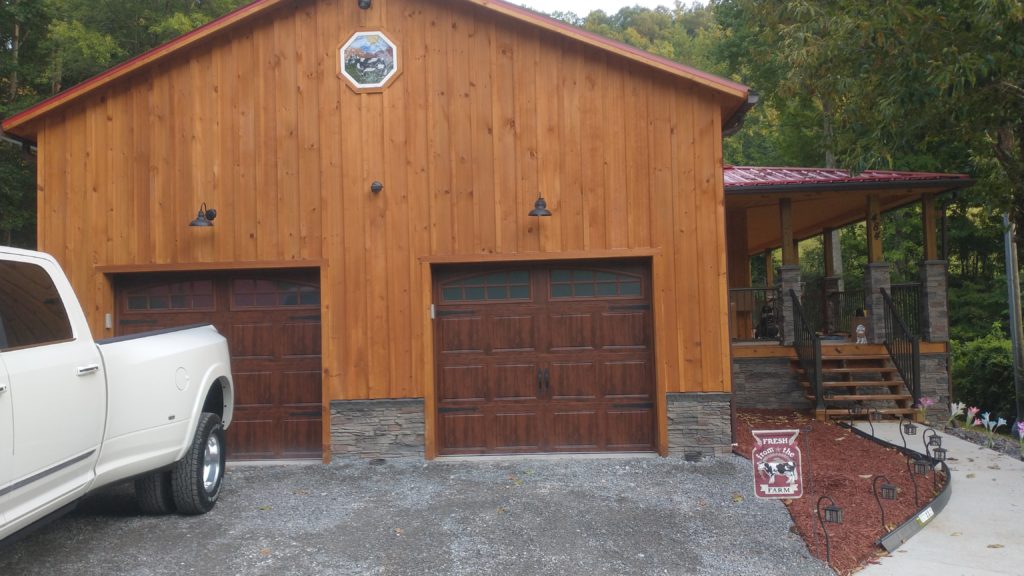 An octagonal stained glass window in a white frame, showcasing a detailed farm scene with three cows—one black and white, two brown—grazing in a green field under a blue sky with distant mountains. The window is installed in a light wood-paneled barn-style building above two dark wood garage doors.