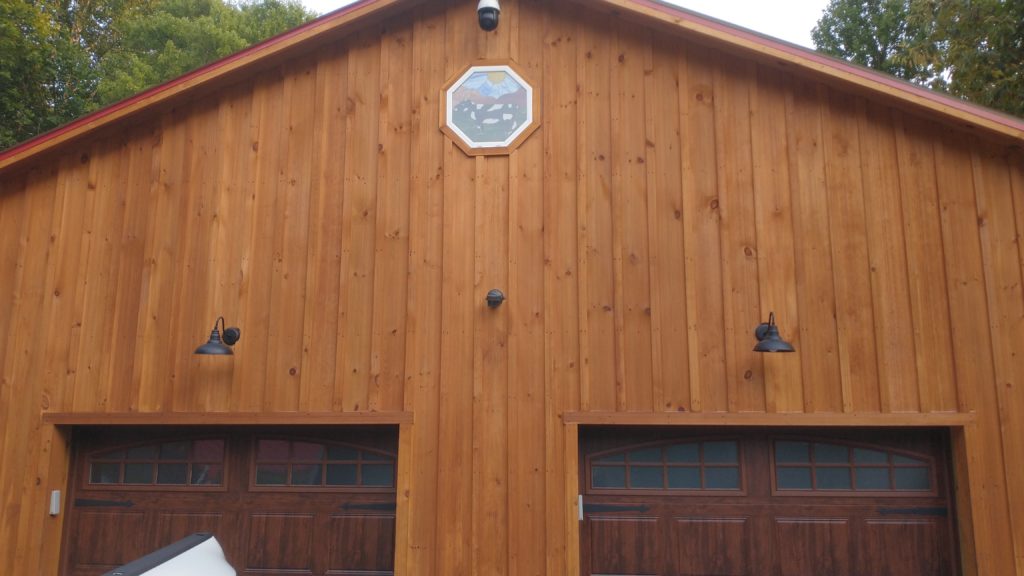 An octagonal stained glass window depicting two black and white cows grazing on green rolling hills under a blue sky with a radiant yellow sun, installed high on a wooden barn gable.