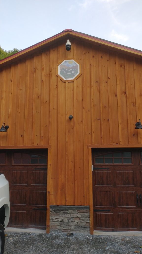 An octagonal stained glass window installed in a light brown wooden barn wall. The glass depicts a pastoral scene with green rolling hills, blue mountains in the background, a herd of white and grey cows in the foreground, a bright yellow sun, and a light blue sky. The window is centered above two dark brown garage doors.