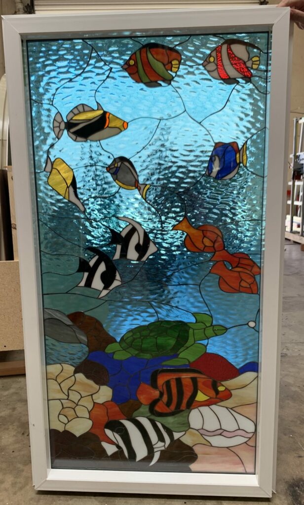 A rectangular stained glass window panel in a white frame, depicting a vibrant underwater scene. Numerous colorful tropical fish, including clownfish and angel fish, a green sea turtle, coral, and seashells are set against a textured blue water background and a seabed of varied colored glass.