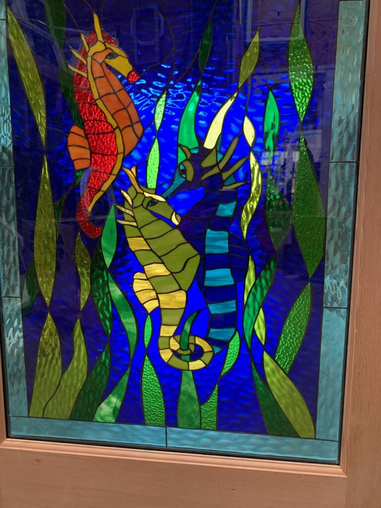 A vibrant rectangular stained glass panel featuring a red-orange seahorse and a green-yellow seahorse amidst flowing green and yellow textured seaweed. The deep blue background glass has a ripple effect, creating an underwater illusion. The panel is set in a light wooden frame.