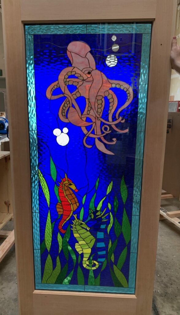 A rectangular stained glass door insert depicting a vibrant underwater scene. A large, pink-orange octopus with visible suction cups on its tentacles is at the top, while a reddish-orange seahorse and a yellow-green seahorse are nestled among varied green seaweed at the bottom. The background is a deep blue textured glass with white bubbles, one of which is distinctly shaped like Mickey Mouse. The entire panel is enclosed in a light wooden frame.