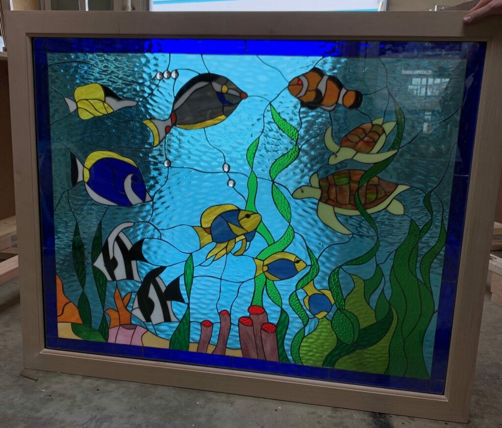 A rectangular stained glass panel in a light wooden frame depicting a vibrant underwater coral reef. Colorful fish, including a clownfish, angelfish, and surgeonfish, swim alongside two sea turtles among green seaweed and red coral. The background is textured light blue glass representing ocean water.
