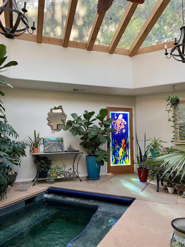 A rectangular stained glass panel set within a light wood door, depicting a vibrant underwater marine life scene. A large, detailed pink octopus with curving tentacles is prominent in the upper section against a rich blue background. Below the octopus, a bright red seahorse and a yellow and blue striped seahorse swim amidst green, wavy seaweed. Small white bubbles rise from the bottom.