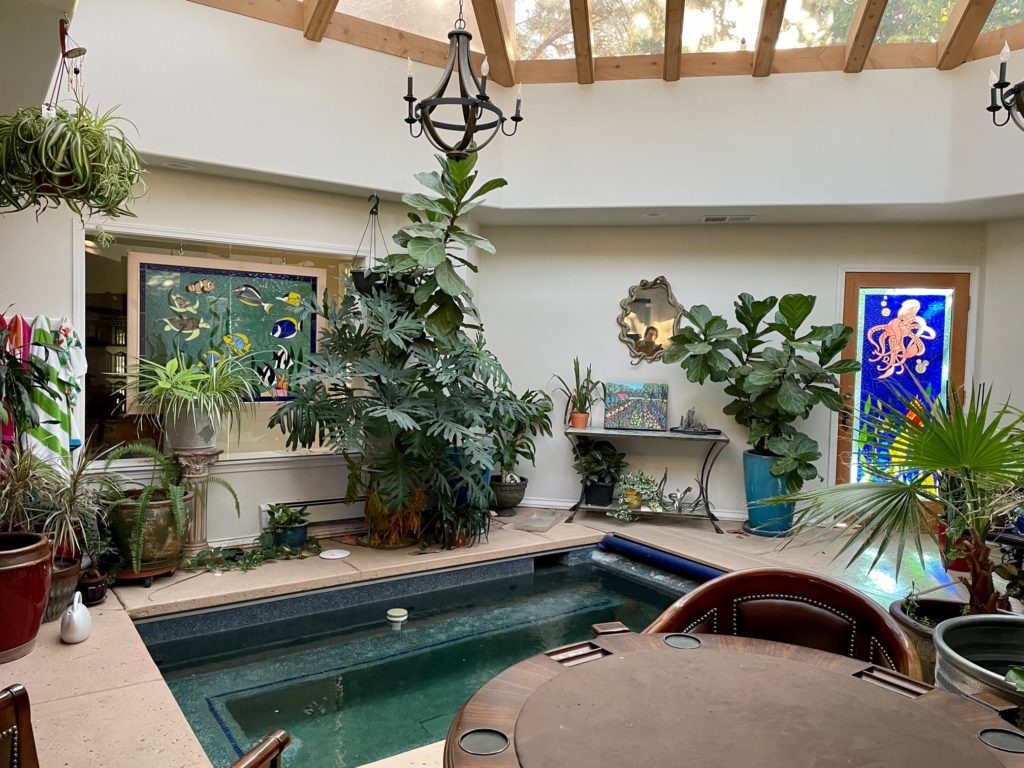 A rectangular stained glass panel depicting a vibrant underwater scene with colorful tropical fish and green sea turtles swimming amidst blue and green textured glass, framed in wood, installed above an interior doorway in a bright sunroom with an indoor pool. A separate stained glass panel with a pink octopus is visible in a door on the right.