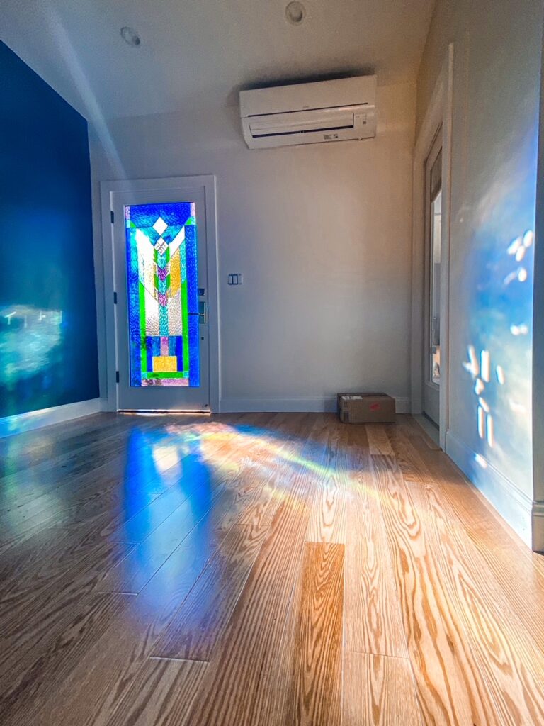 A tall white interior door featuring a vivid, geometric stained glass panel. The panel showcases abstract patterns in various shades of blue, green, and yellow glass. Sunlight streams through the glass, projecting colorful, rainbow-like refractions onto a light hardwood floor and an adjacent white wall in a modern room with a dark blue accent wall.