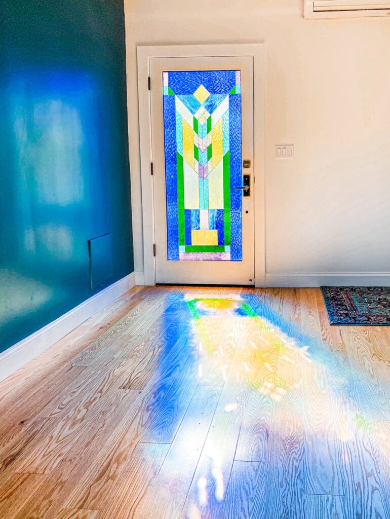 A full-light white entry door featuring a vibrant, leaded stained glass panel in a geometric style, inspired by Art Deco or Craftsman design. The panel prominently features rectangular and triangular shapes in various shades of blue, yellow, green, and clear textured glass. Sunlight streams through the glass, casting colorful, abstract light patterns onto the light wooden floor, illuminating a nearby teal wall.
