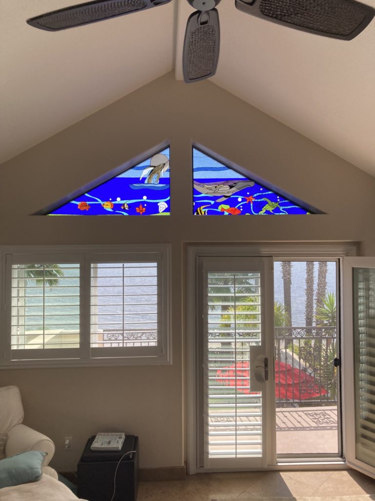 Two custom triangular stained glass window panels installed in the gables of a vaulted ceiling, depicting a vibrant underwater scene. The left panel shows a jellyfish and various fish, while the right panel features a large whale, sea turtles, and more fish, all against a deep blue water background. Below, white-shuttered windows and a sliding door offer a clear view of the ocean.