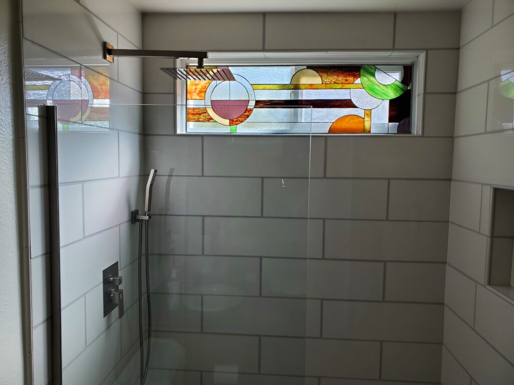 A contemporary bathroom shower with light gray subway tiles, a square shower head, and a glass door, featuring a horizontal leaded stained glass transom window. The abstract geometric design includes circles and horizontal bands in amber, orange, green, and clear textured glass.