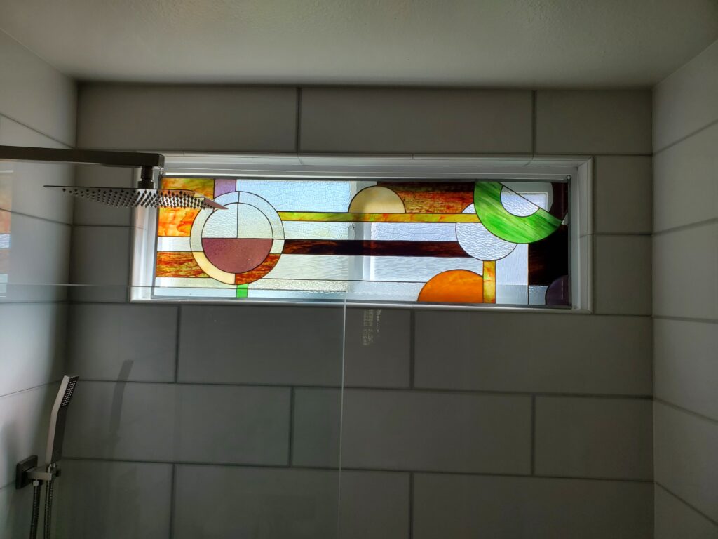 A rectangular stained glass window with a contemporary abstract geometric design, installed horizontally above a shower. It features intersecting circles and straight lines in shades of warm orange, yellow, maroon, vibrant green, and various clear textured glass panels, set within a white frame against light gray tiled walls.