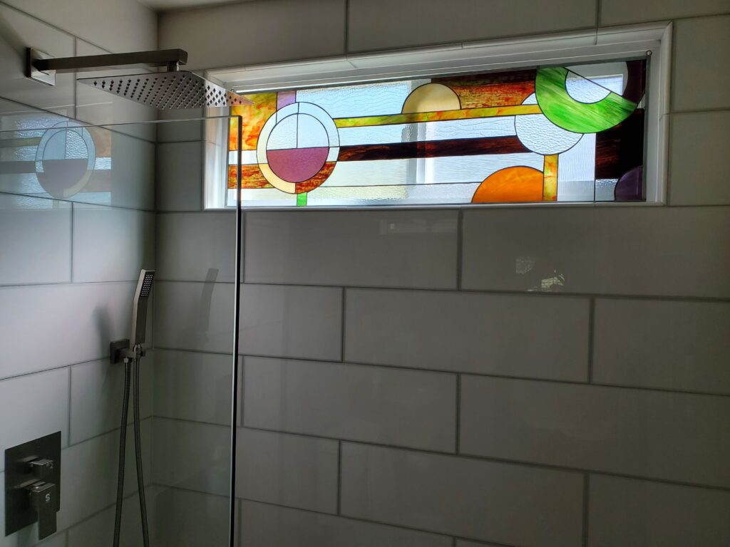 A rectangular, leaded stained glass transom window installed above a modern shower. The abstract geometric design features interlocking circles and horizontal bands in textured clear, amber, green, purple, and light blue glass, framed by white subway tiles and reflecting light from a showerhead.