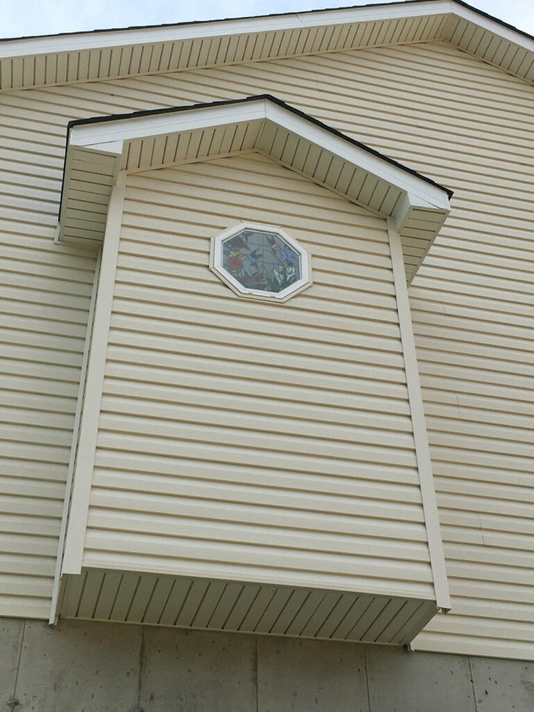 An octagonal stained glass window depicting hummingbirds and colorful flowers, installed in a beige vinyl-sided dormer on a house exterior.