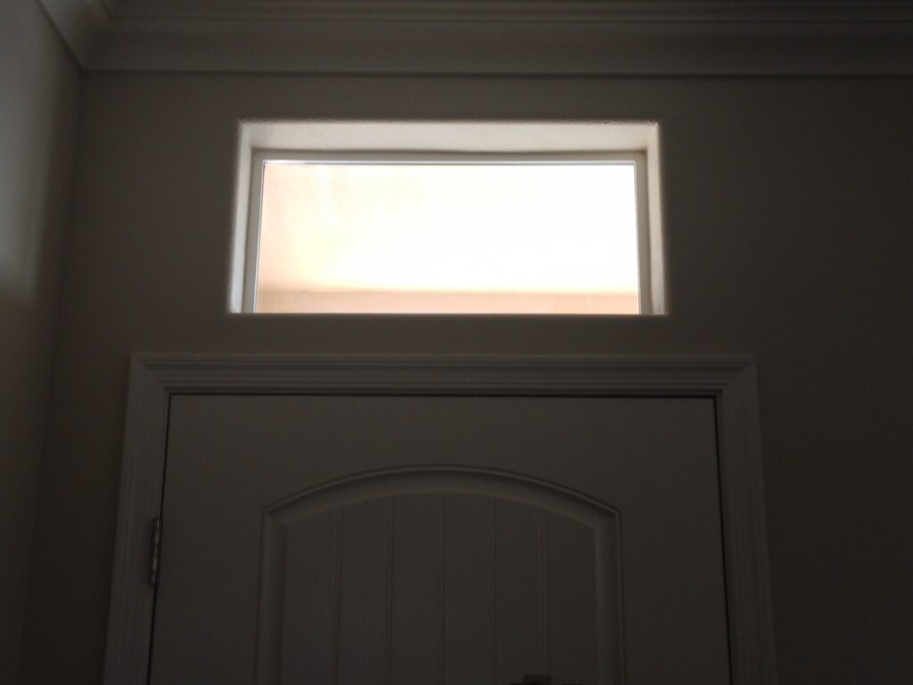 A rectangular transom window with frosted or obscure glass installed above a white paneled door, allowing diffused light into a dimly lit room.