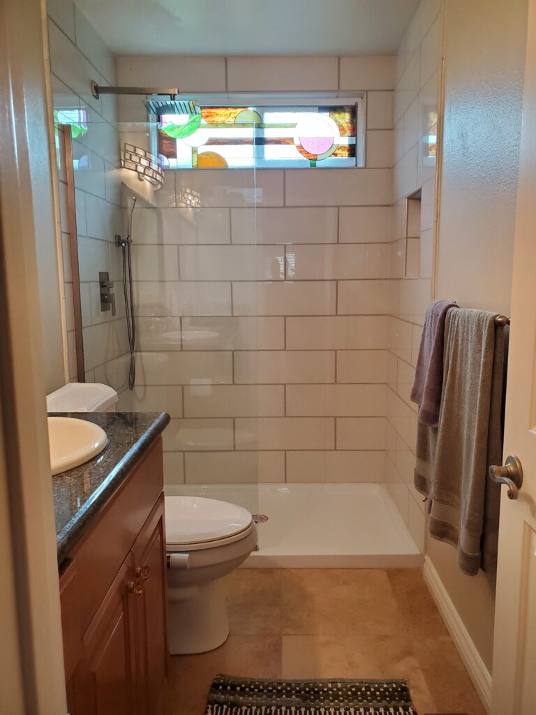 A horizontal stained glass transom window with a contemporary abstract geometric design, featuring circles, rectangles, and curved shapes in clear, green, orange, brown, and pink art glass. The window is positioned above a white tiled shower enclosure in a modern bathroom.