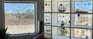 A composite image displaying two contrasting stained glass installations. On the left, a large rectangular window features a leaded clear glass panel with a complex abstract geometric design created from various sizes of clear beveled and textured rectangles, providing privacy and diffusing natural light. On the right, a built-in shelving unit houses multiple square stained glass panels, each adorned with an abstract, whimsical pattern of clear textured glass accented by various shades and sizes of blue and green circles and small squares, connected by intricate lead lines. Decorative objects like a sailboat model are placed on the shelves between the panels.