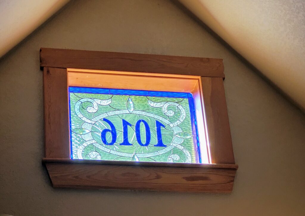 A rectangular leaded stained glass panel in a dark wooden frame, installed in a light brown wall of a sloped attic ceiling. The panel features the word 'EVOL' in bold blue glass, designed to be read as 'LOVE' from the exterior, set against a textured light green background with clear, ornate swirled designs. Bright sunlight filters through the glass, illuminating its vibrant colors.