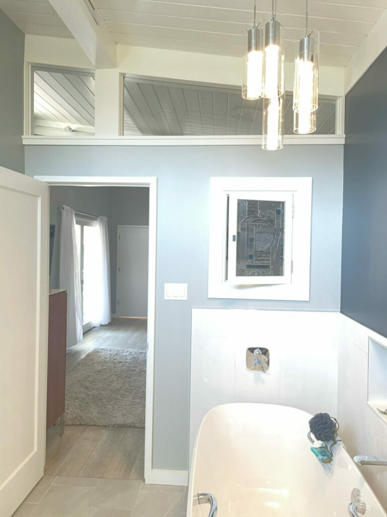 A rectangular leaded stained glass window featuring an abstract geometric pattern with clear textured glass and small blue square accents, installed in a white frame above a white bathtub in a modern light gray and white bathroom.