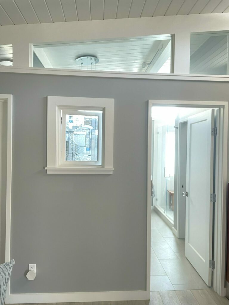 A small square window with a leaded glass panel, featuring a modern geometric design with clear textured glass, clear beveled elements, and subtle light blue accent pieces. The panel is set within a white frame on a light gray wall.