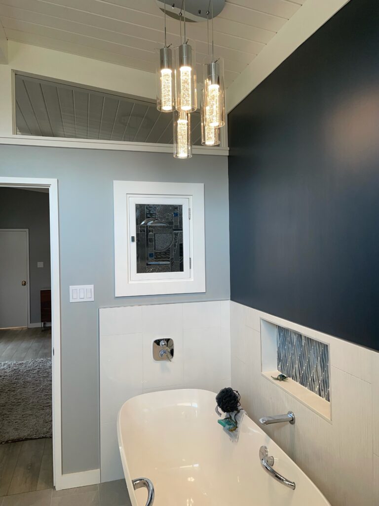 A modern bathroom showcases a recessed rectangular stained glass panel featuring an abstract geometric design crafted from clear textured and beveled glass with subtle blue accents, mounted above a white freestanding bathtub.