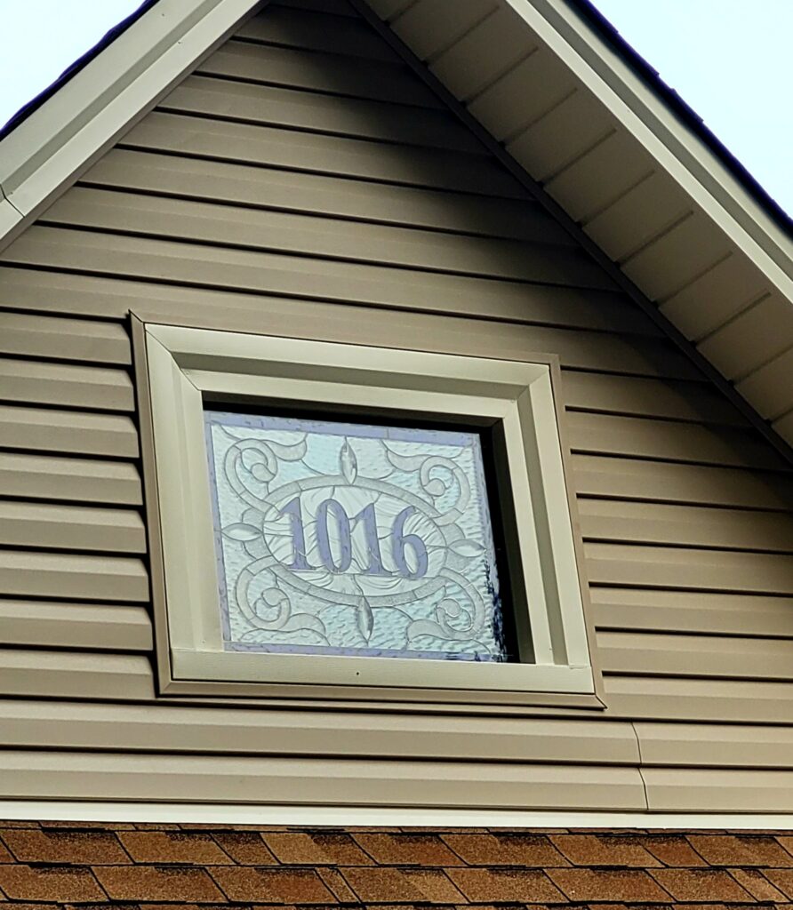A rectangular leaded stained glass panel displaying the house number '1016' in light purple glass, surrounded by clear textured glass with an elegant swirling design and small bevels, installed in a gable window of a tan vinyl-sided house.
