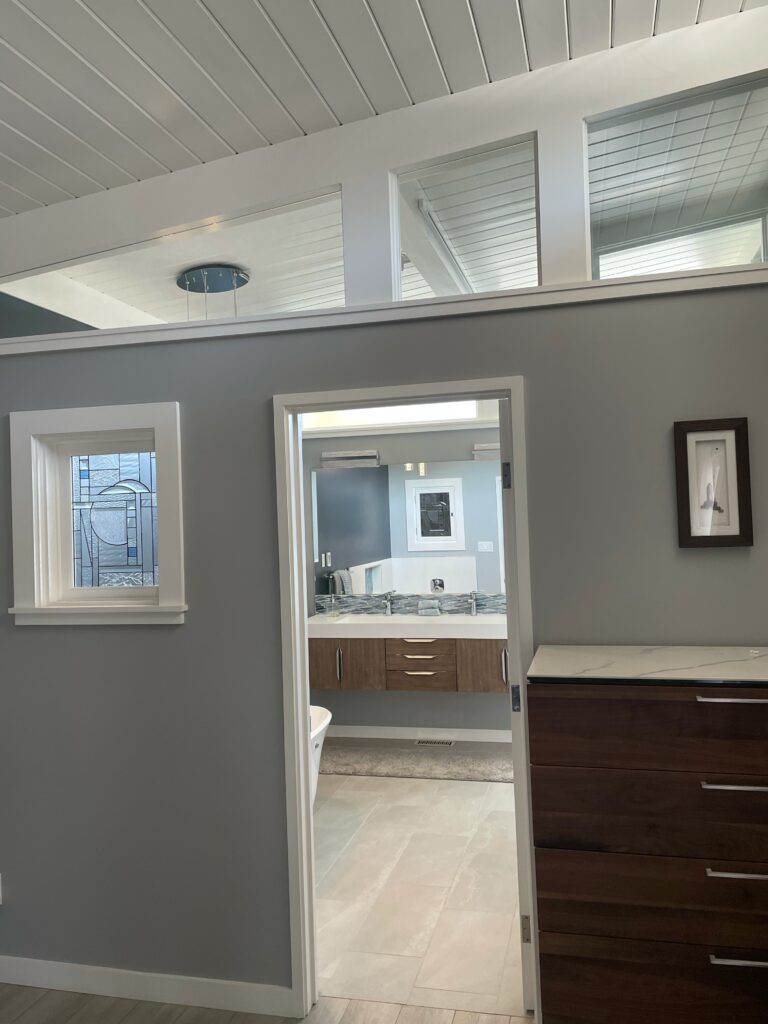 A rectangular stained glass window featuring a modern abstract geometric design with various clear textured glass panels and accent pieces of light blue and grey glass, set in a white frame on a grey wall.