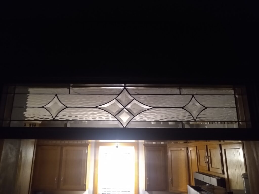 A rectangular leaded stained glass transom panel featuring a central geometric design of clear beveled diamonds and curved shapes, flanked by sections of clear ripple-textured glass. The panel is installed above an interior archway, with a brightly lit kitchen visible below.