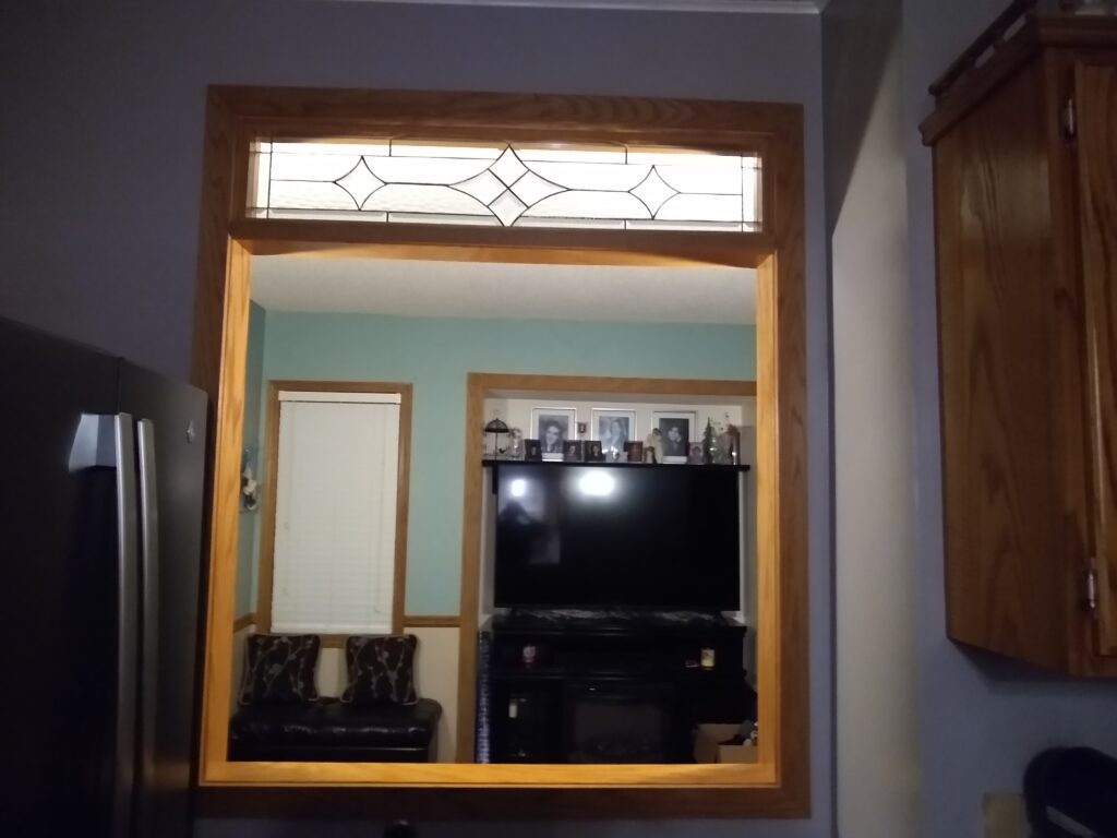 A rectangular clear textured stained glass transom panel with a modern geometric leaded design featuring elongated diamonds, set in a light wood frame above an interior room opening.