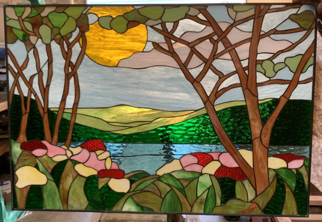 A rectangular stained glass panel depicting a vibrant, serene landscape. The foreground features lush green foliage with large pink and yellow flowers and textured red details. A calm blue lake occupies the midground, bordered by rolling green hills in the background under a light blue sky. A prominent, textured yellow sun shines in the upper center, while several trees with detailed brown branches and light green leaves frame the scene.