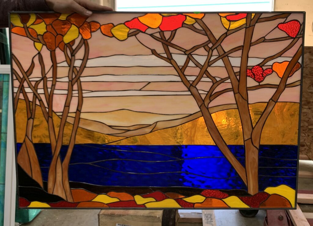 A rectangular stained glass panel featuring a vibrant autumnal landscape. Two prominent brown trees with intricate branches and bright red, orange, and yellow leaves frame a deep blue lake. A golden-orange shore or distant hills rise behind the water, while the sky above displays soft horizontal bands of pale pink and orange. The foreground shows abstract shapes of red, orange, and yellow, resembling fallen leaves.
