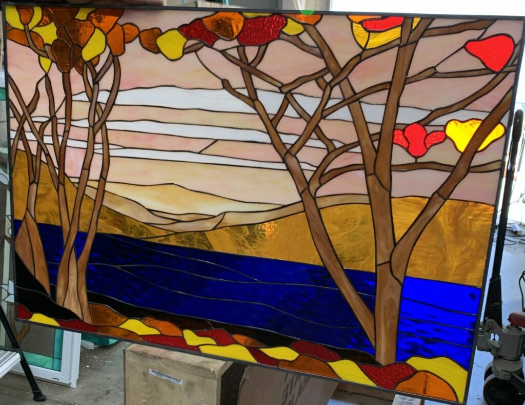 A rectangular leaded stained glass panel depicting a stylized autumn landscape. The foreground features bare trees with vibrant red, orange, and yellow leaves. A deep blue river flows horizontally through the mid-ground, while golden and ochre hills rise in the background under a layered sky of soft pinks, peaches, and whites.